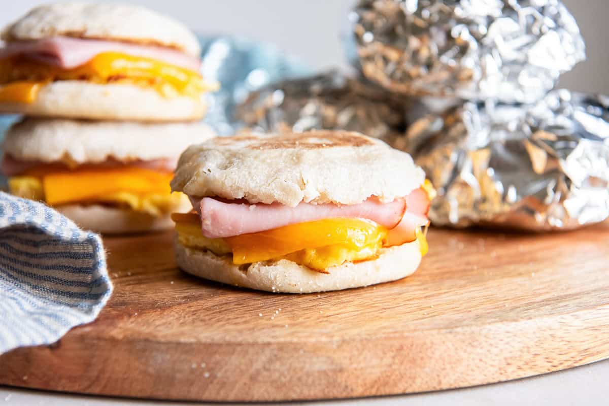 Ham, egg, and melted cheddar breakfast sandwich on an English muffin, with more sandwiches and foil-wrapped sandwiches in the background.