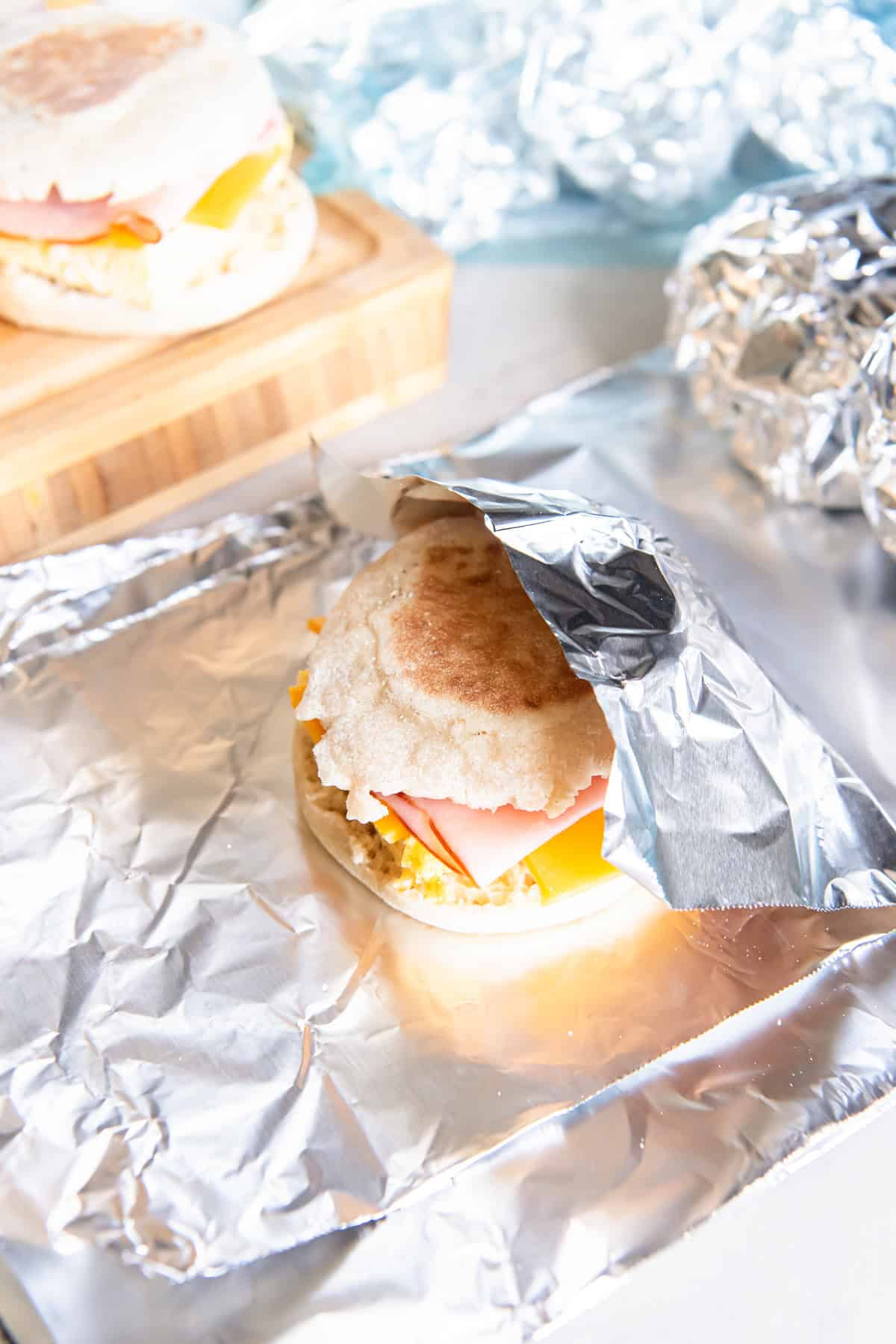 Ham, egg, and cheese breakfast sandwich partially wrapped in foil, with additional wrapped sandwiches in the background.