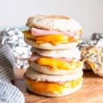 Three ham, egg, and cheese breakfast sandwiches stacked on a wooden board with foil-wrapped sandwiches in the background.
