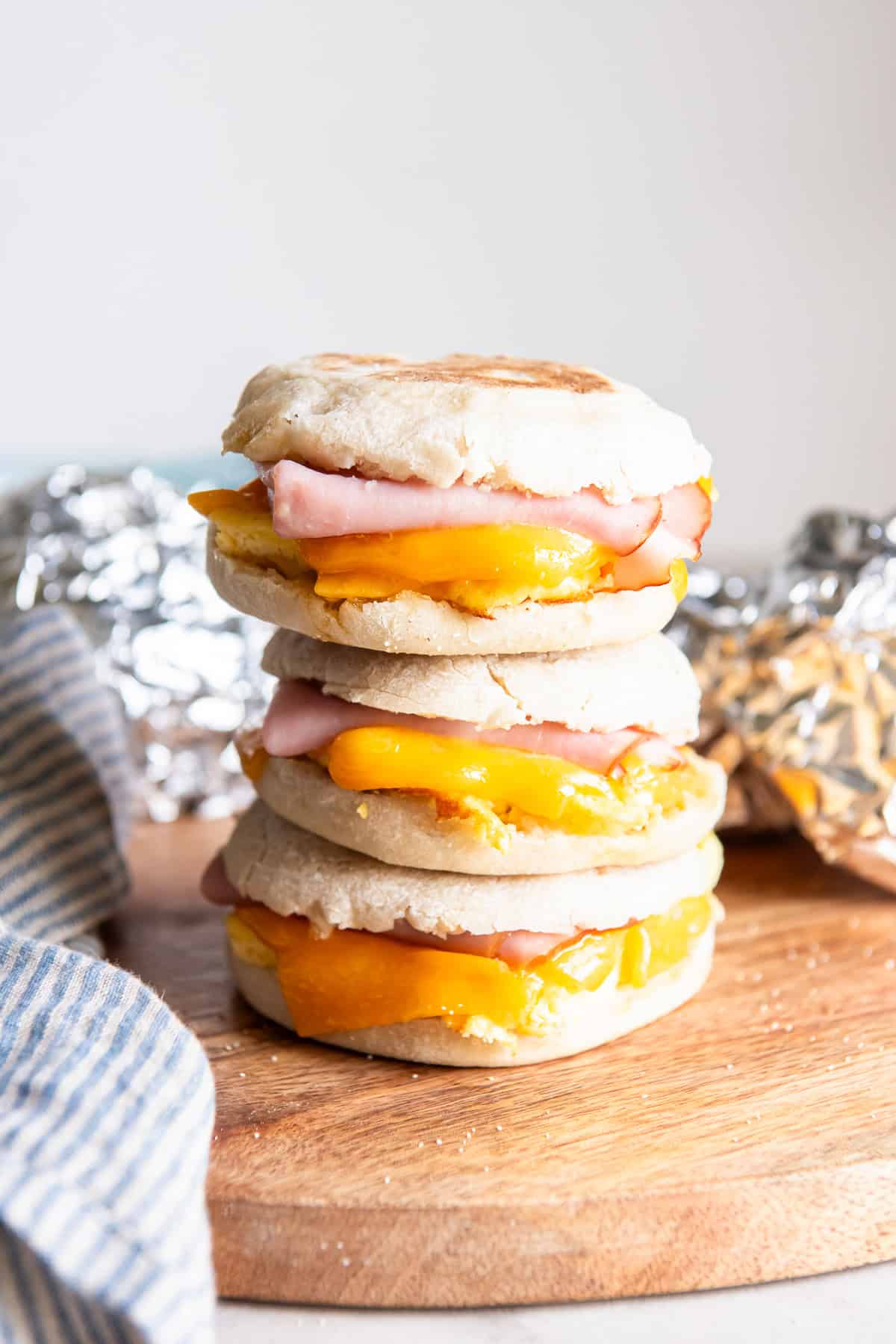 Three ham, egg, and cheese breakfast sandwiches stacked on a wooden board with foil-wrapped sandwiches in the background.