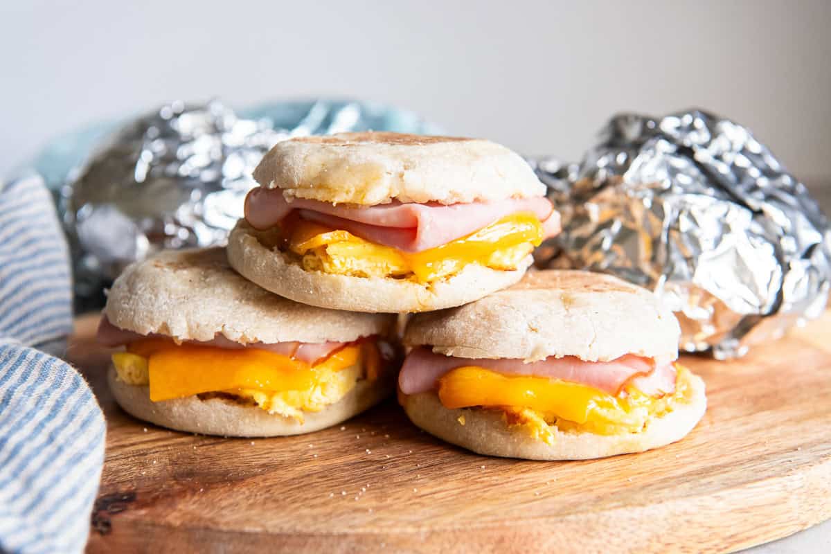 Three ham, egg, and cheese breakfast sandwiches on a wooden board with foil-wrapped sandwiches in the background.