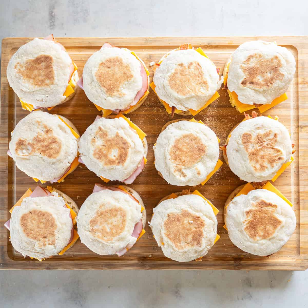 Twelve assembled ham and bacon breakfast sandwiches on English muffins, ready to wrap and store.