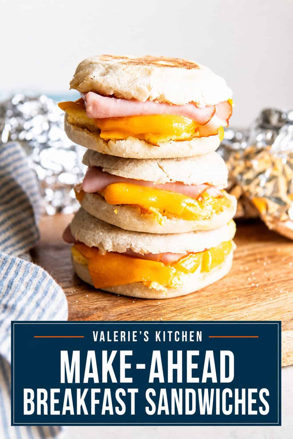 Three ham, egg, and cheese breakfast sandwiches stacked on a wooden board with foil-wrapped sandwiches in the background with bold title text overlay.