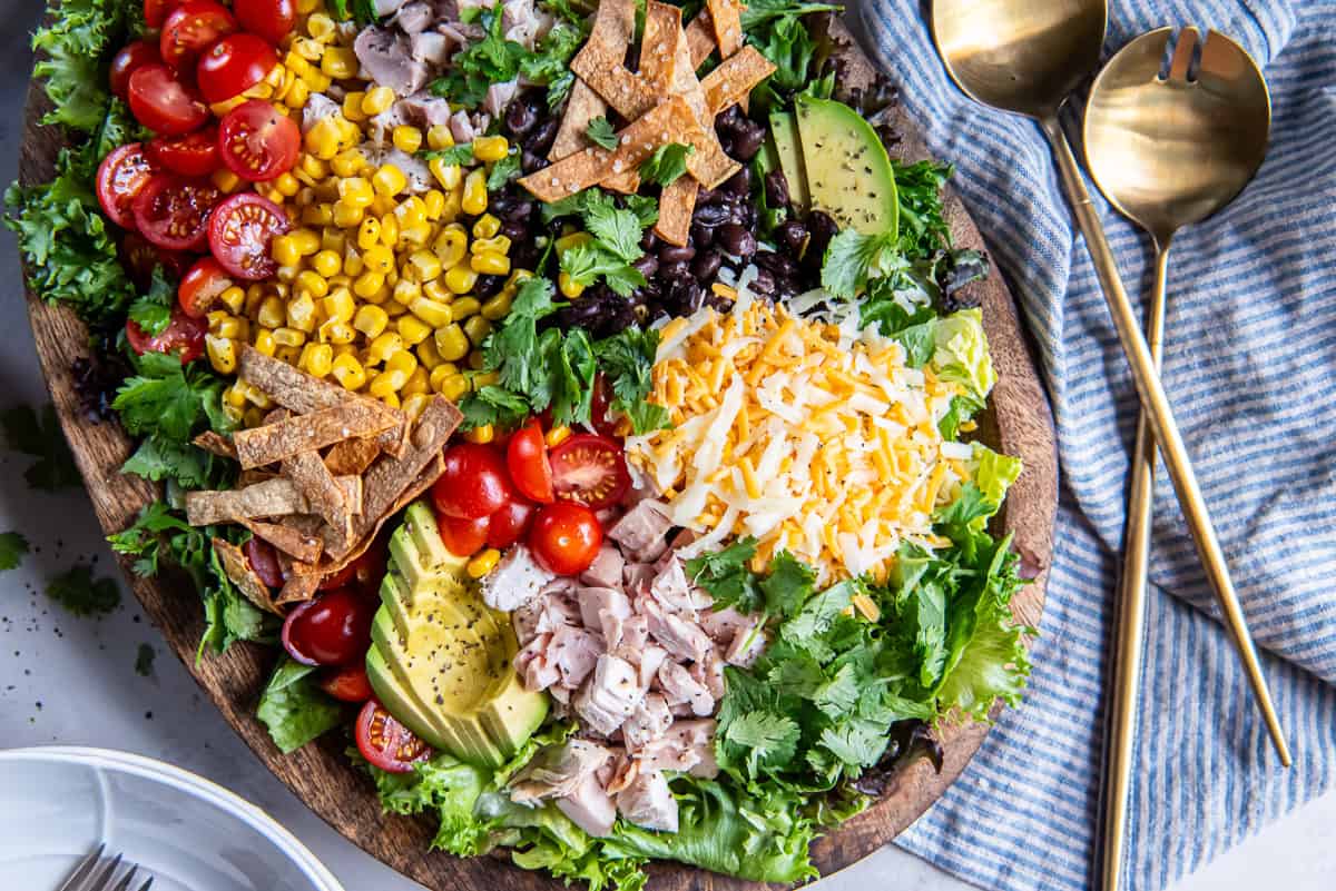 An assembled Southwest Chicken Caesar Salad with romaine lettuce, chicken, avocado, black beans, corn, tomatoes, shredded cheese, fresh cilantro, and crispy tortilla strips before adding Caesar dressing.