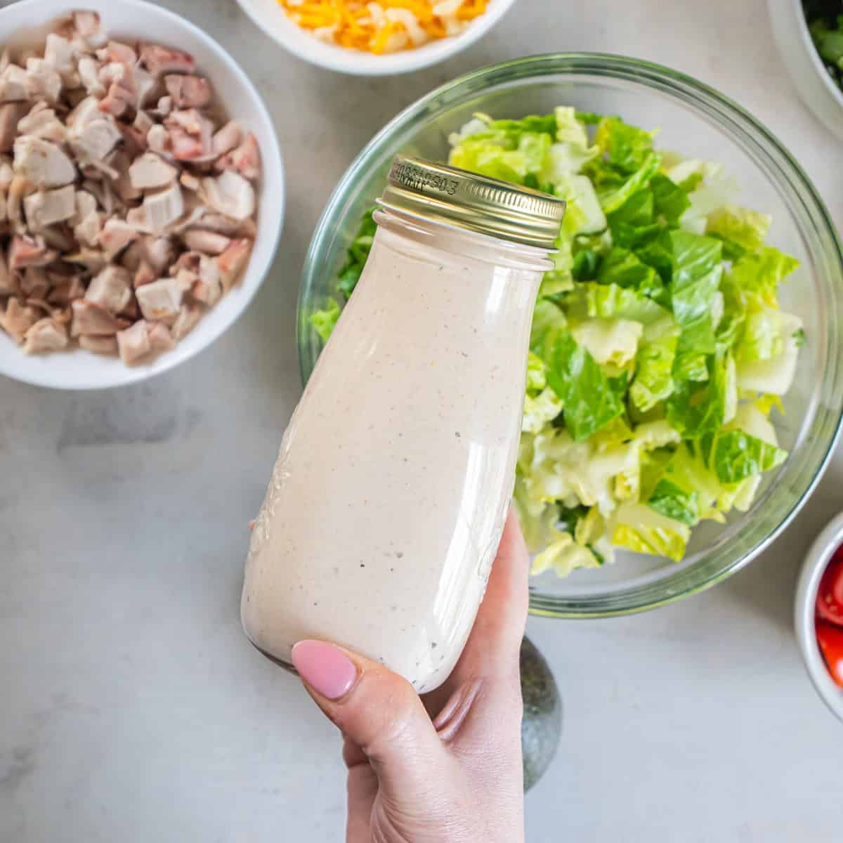 A bottle of creamy Southwest Caesar dressing held over a bowl of chopped romaine with the salad ingredients in the background.