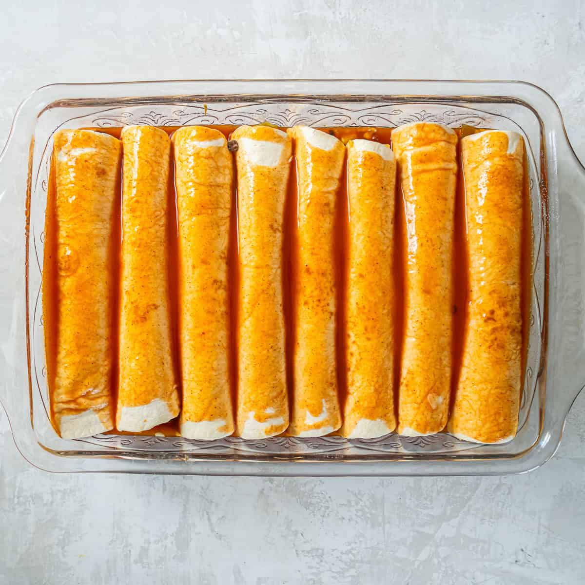 Eight rolled chicken enchiladas arranged seam-side down in a baking dish and covered with red enchilada sauce.