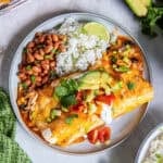 Easy Southwest Chicken Enchiladas topped with avocado, sour cream, tomatoes, green onions, and cilantro, served with rice and pinto beans.