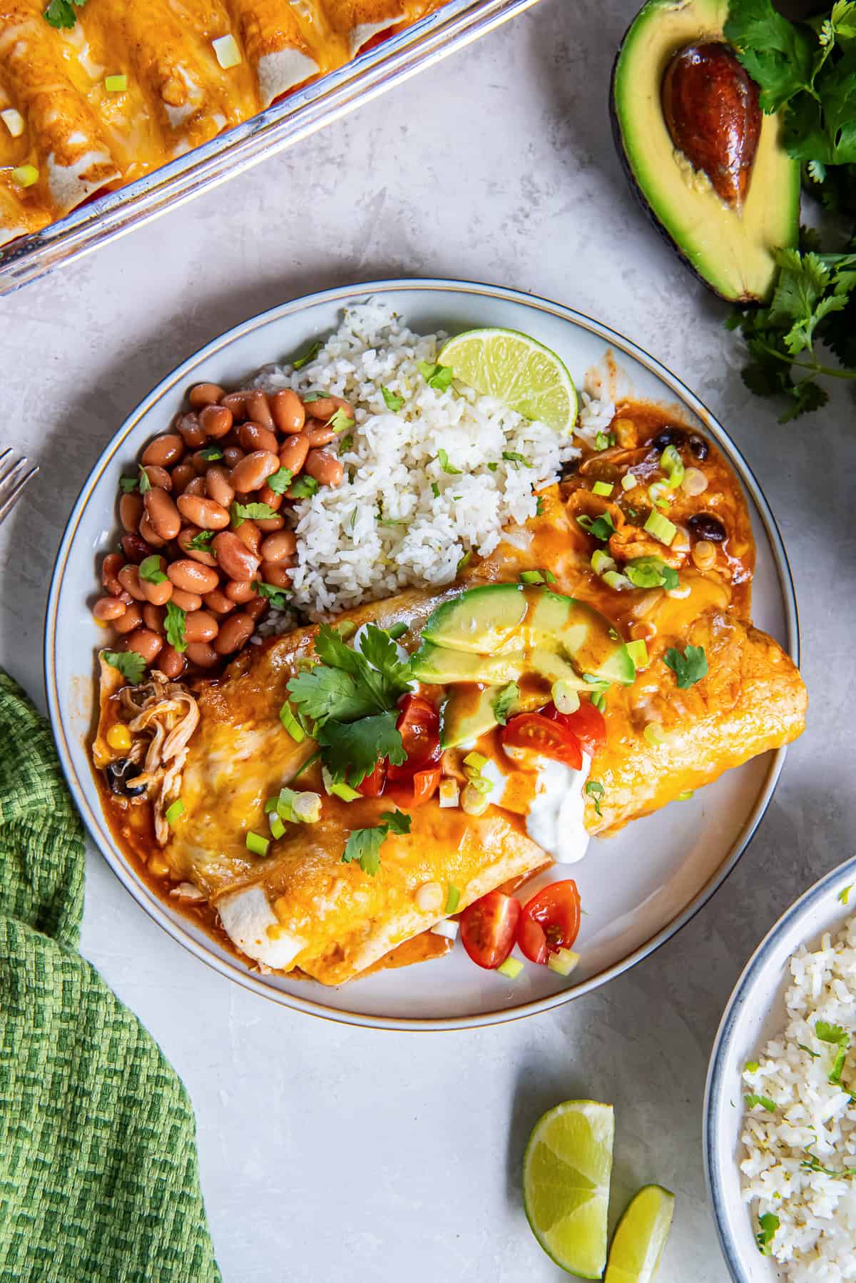 Easy Southwest Chicken Enchiladas topped with avocado, sour cream, tomatoes, green onions, and cilantro, served with rice and pinto beans.