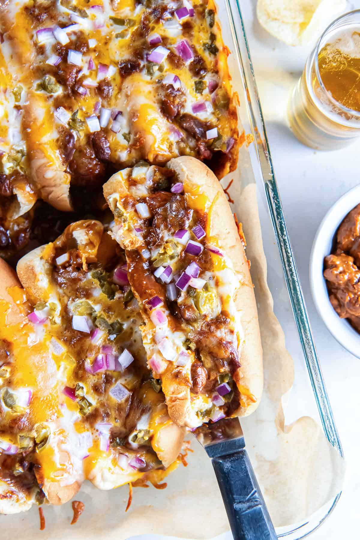 A baked chili dog being lifted from a pan of chili dogs topped with melted cheese, red onion, and pickled jalape&ntilde;os.