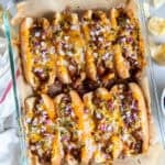 Eight baked chili dogs topped with melted cheese, red onion, and pickled jalapeños in a baking dish with beer and potato chips.