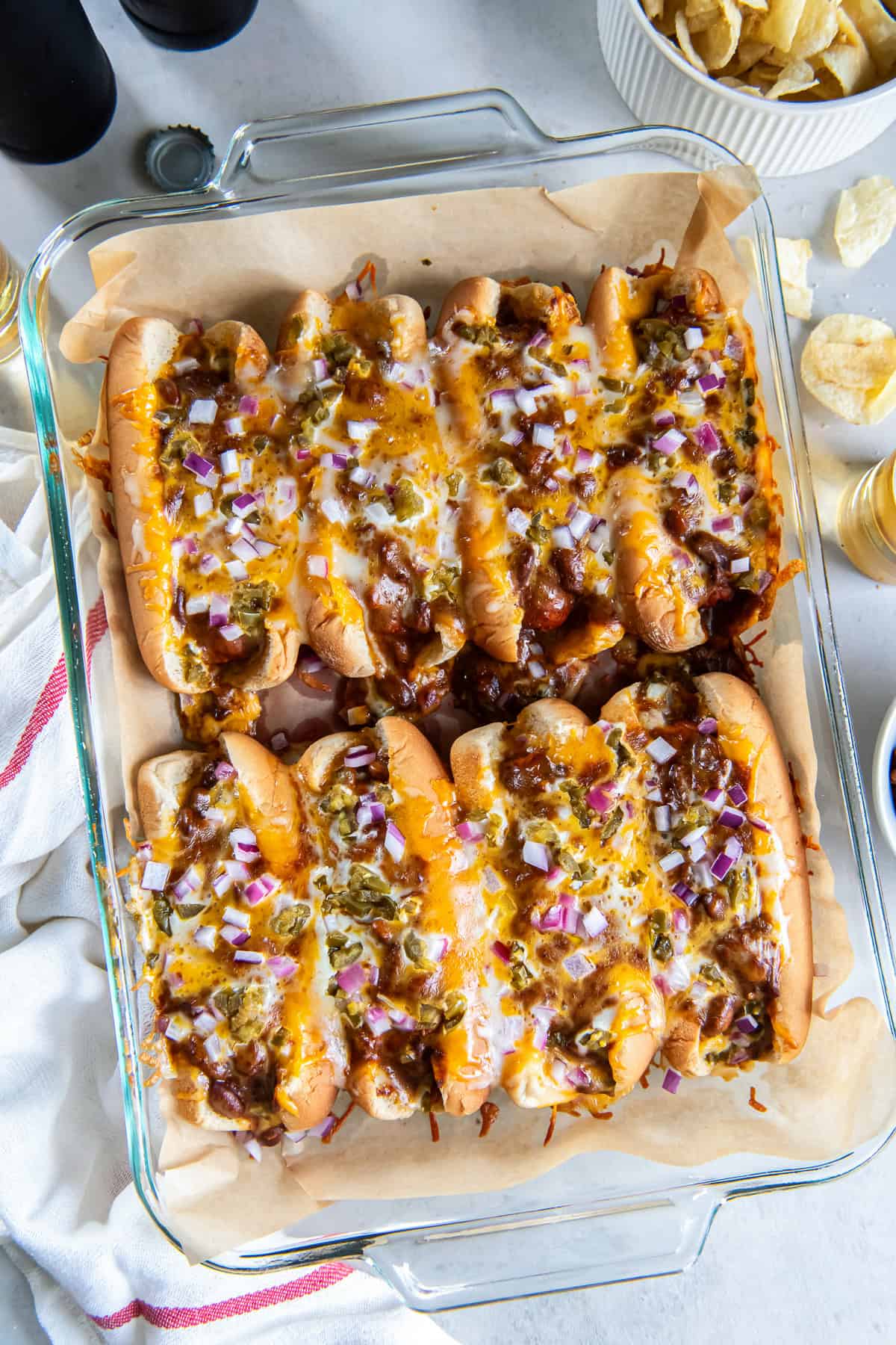 Eight baked chili dogs topped with melted cheese, red onion, and pickled jalape&ntilde;os in a baking dish with beer and potato chips.