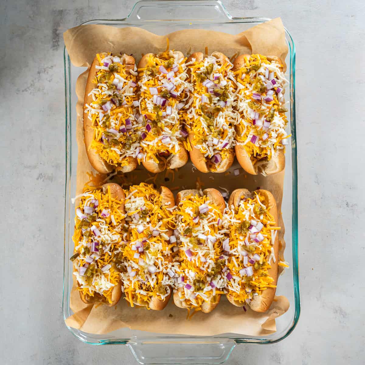 Baked chili dogs with melted cheese, diced red onion, and pickled jalape&ntilde;os in a baking dish.