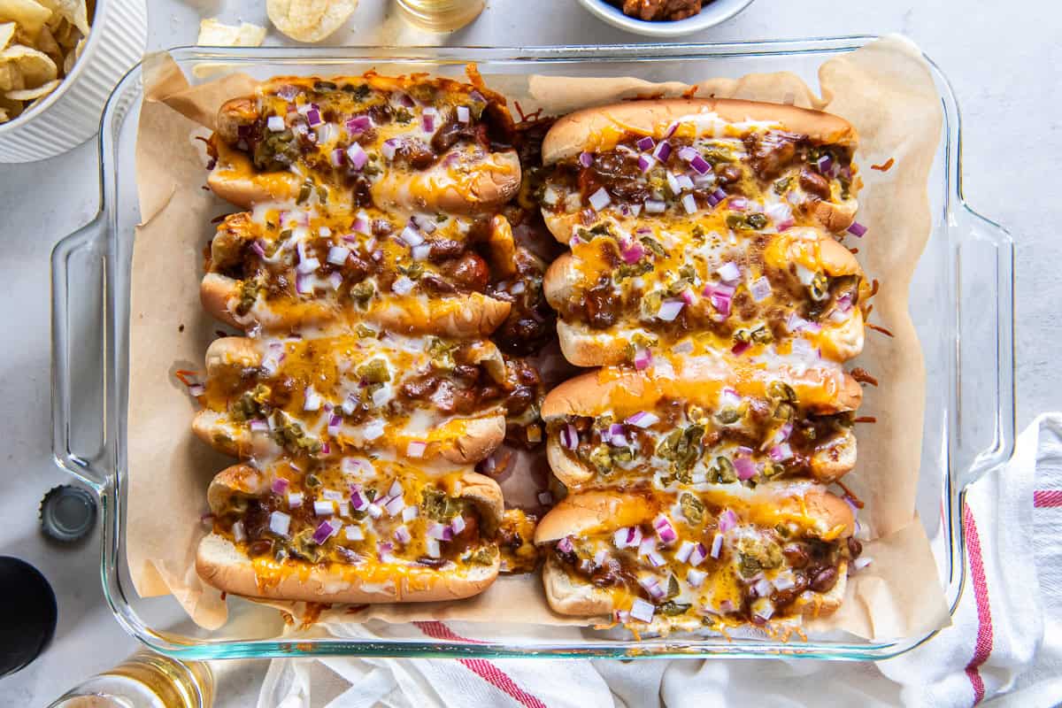 Eight baked chili dogs topped with melted cheese, red onion, and pickled jalape&ntilde;os in a baking dish with beer and potato chips.