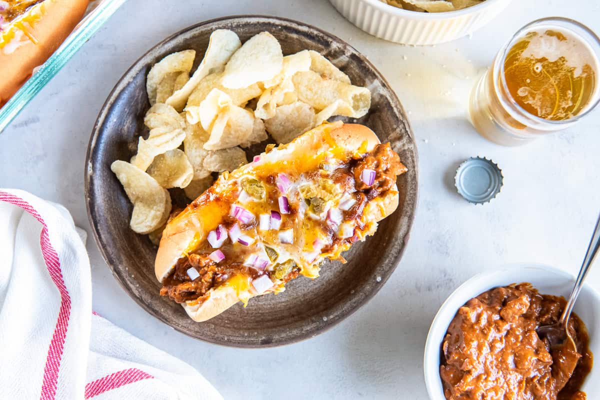 A baked chili dog topped with melted cheese, red onion, and pickled jalape&ntilde;os served with potato chips.