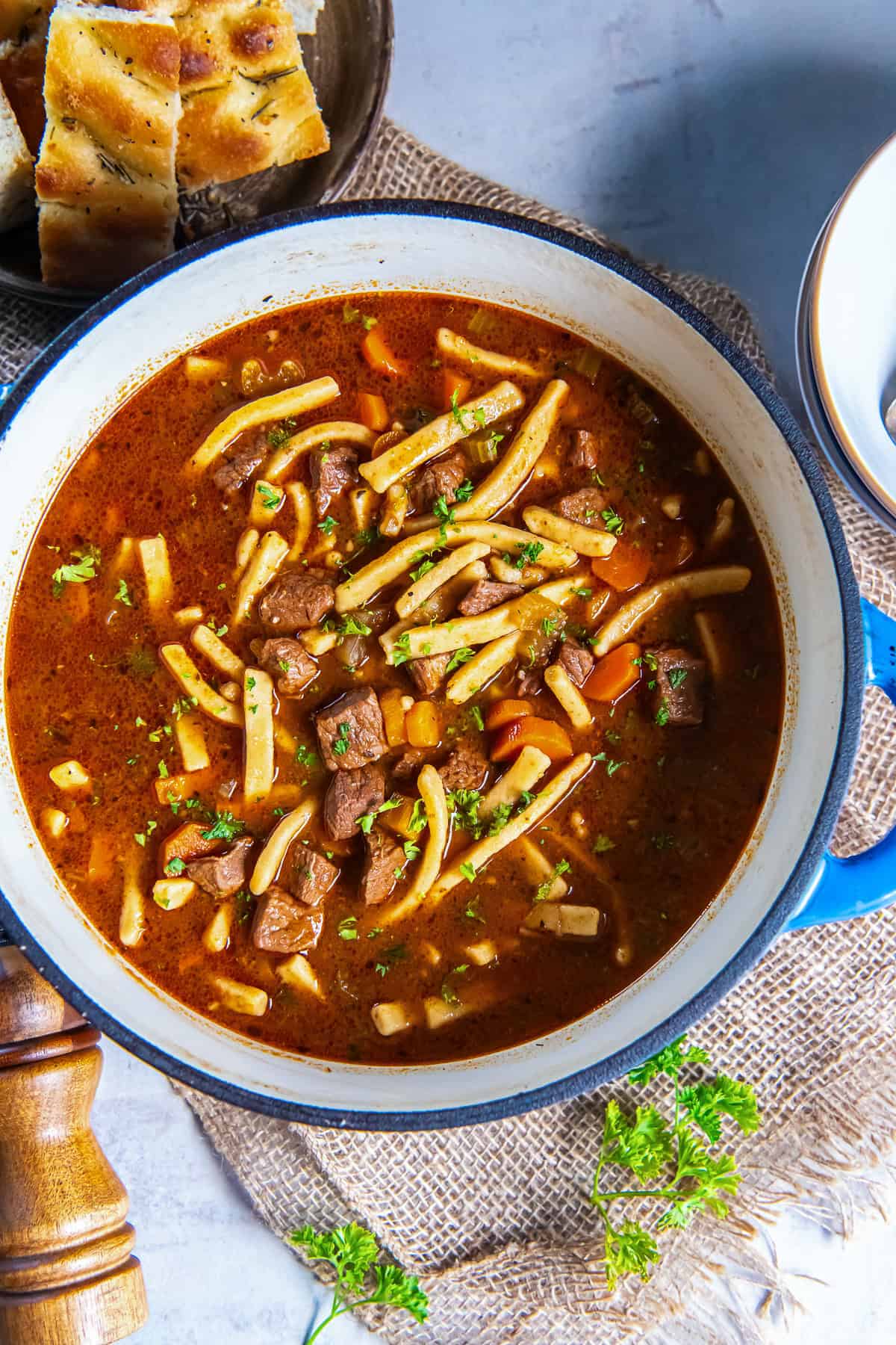 Beef noodle soup with tender chunks of beef, egg noodles, carrots, and celery in a rich broth, served in a blue Dutch oven.