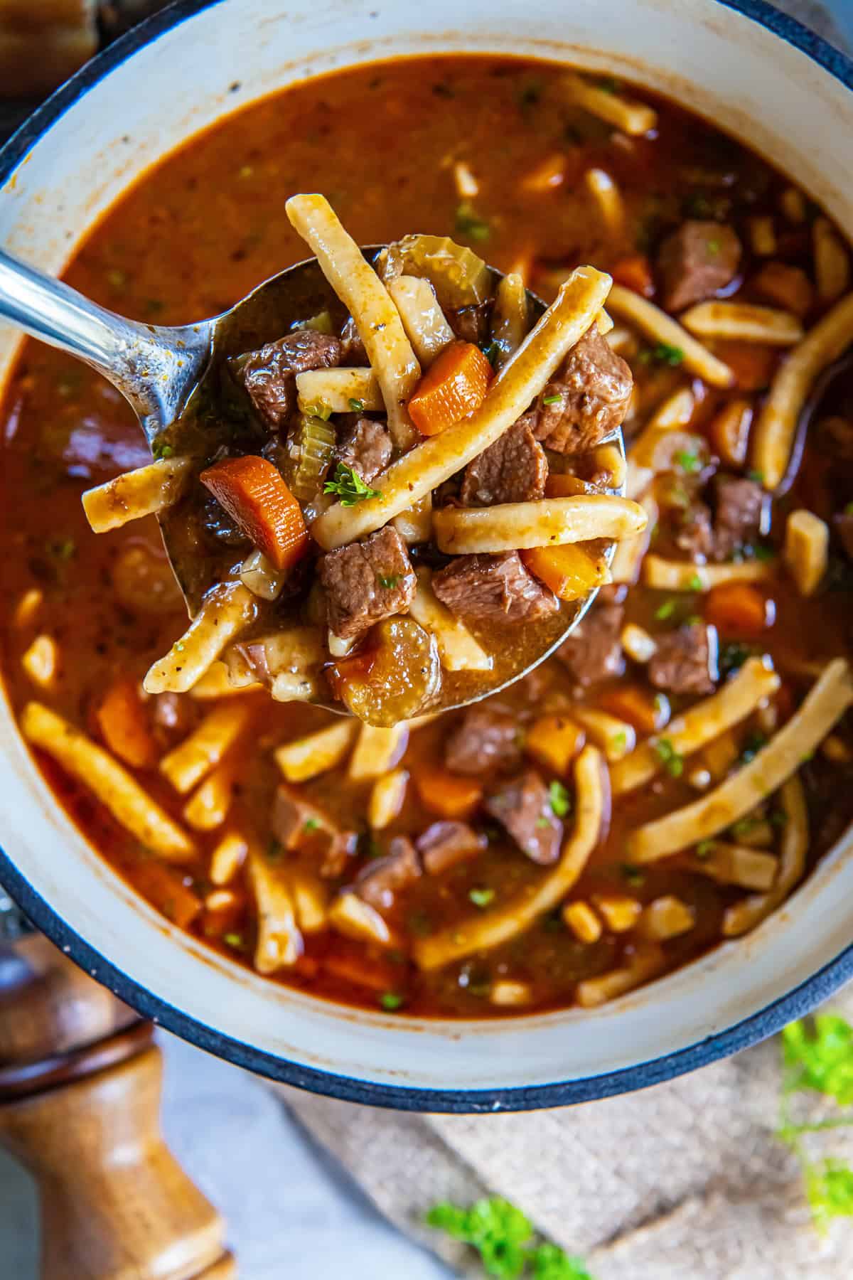 Ladle filled with beef, egg noodles, carrots, and celery over a Dutch oven of homemade beef noodle soup.