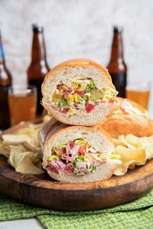 Chopped Italian Sandwich halves stacked on a wooden board, showing the creamy chopped deli meat, cheese, lettuce, tomato, pickles, and banana pepper filling.