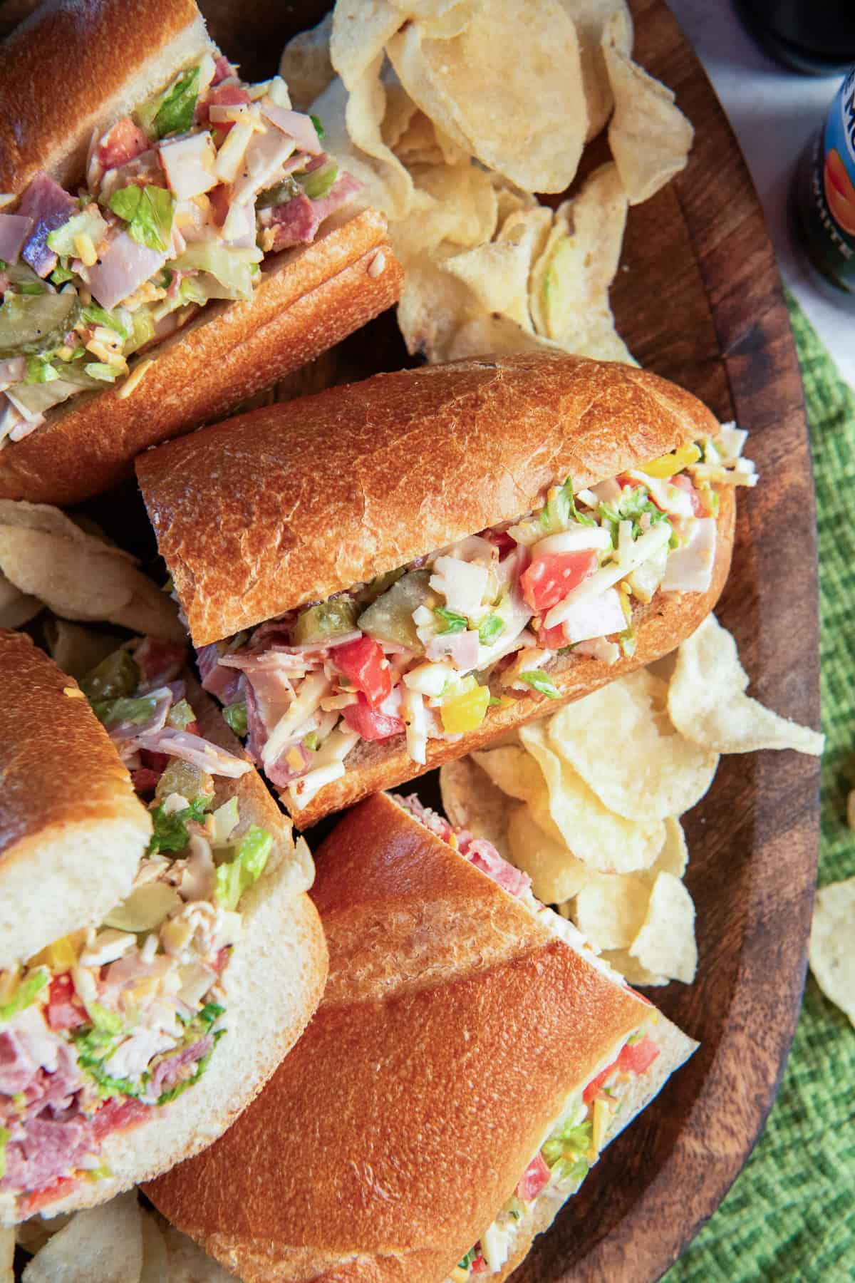 Chopped Italian Sandwich portions filled with deli meats, cheese, lettuce, tomato, pickles, and banana peppers, served with potato chips on a wooden board.