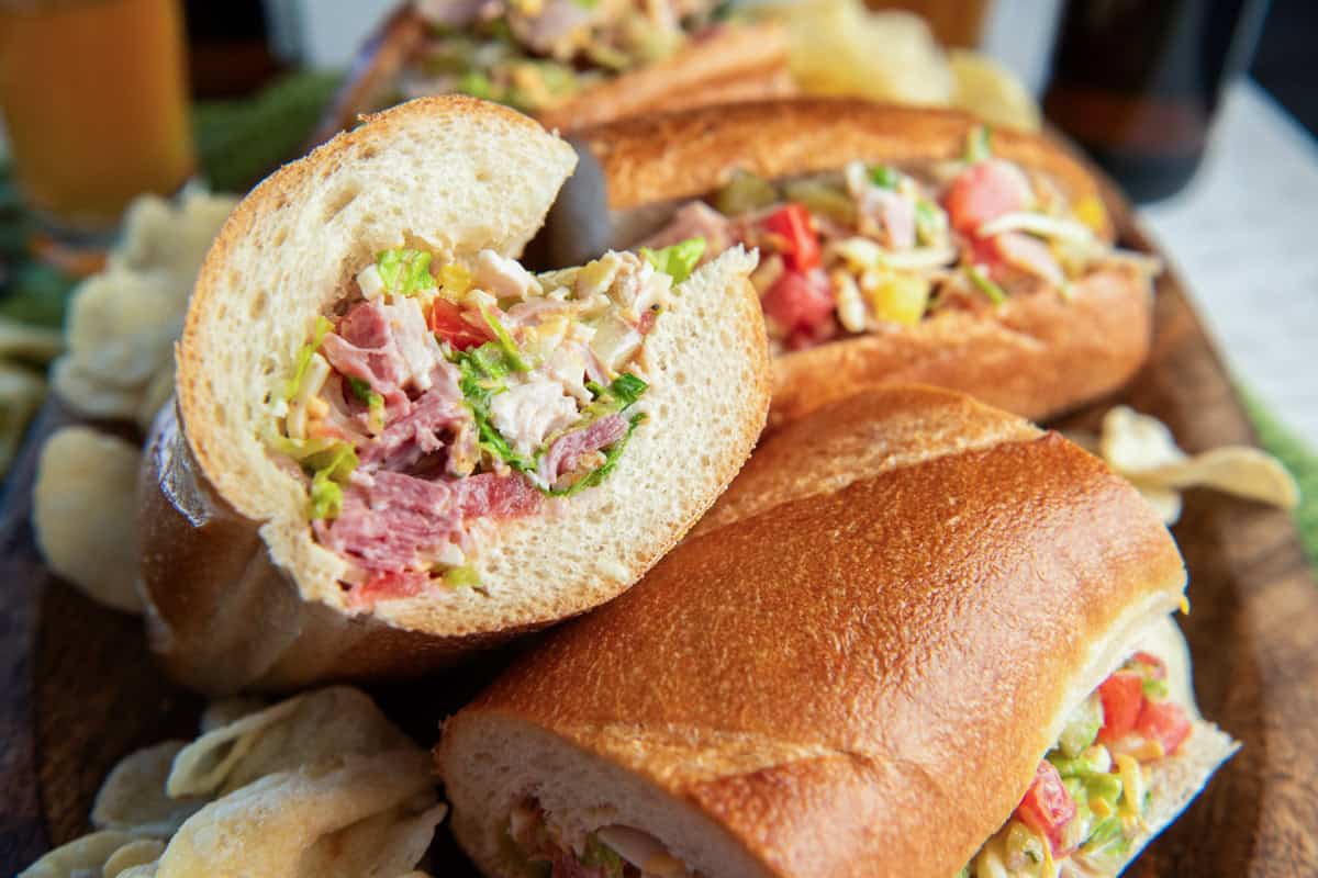 Chopped Italian Sandwiches filled with deli meats, cheese, lettuce, tomato, pickles, and banana peppers, served with potato chips on a wooden board.