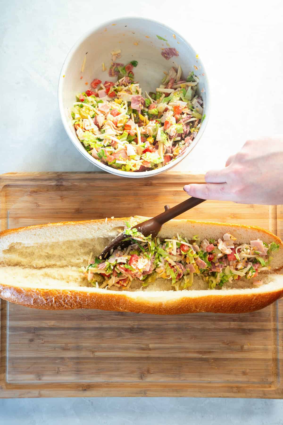 Chopped Italian sandwich filling being spooned into a hollowed French baguette on a cutting board.