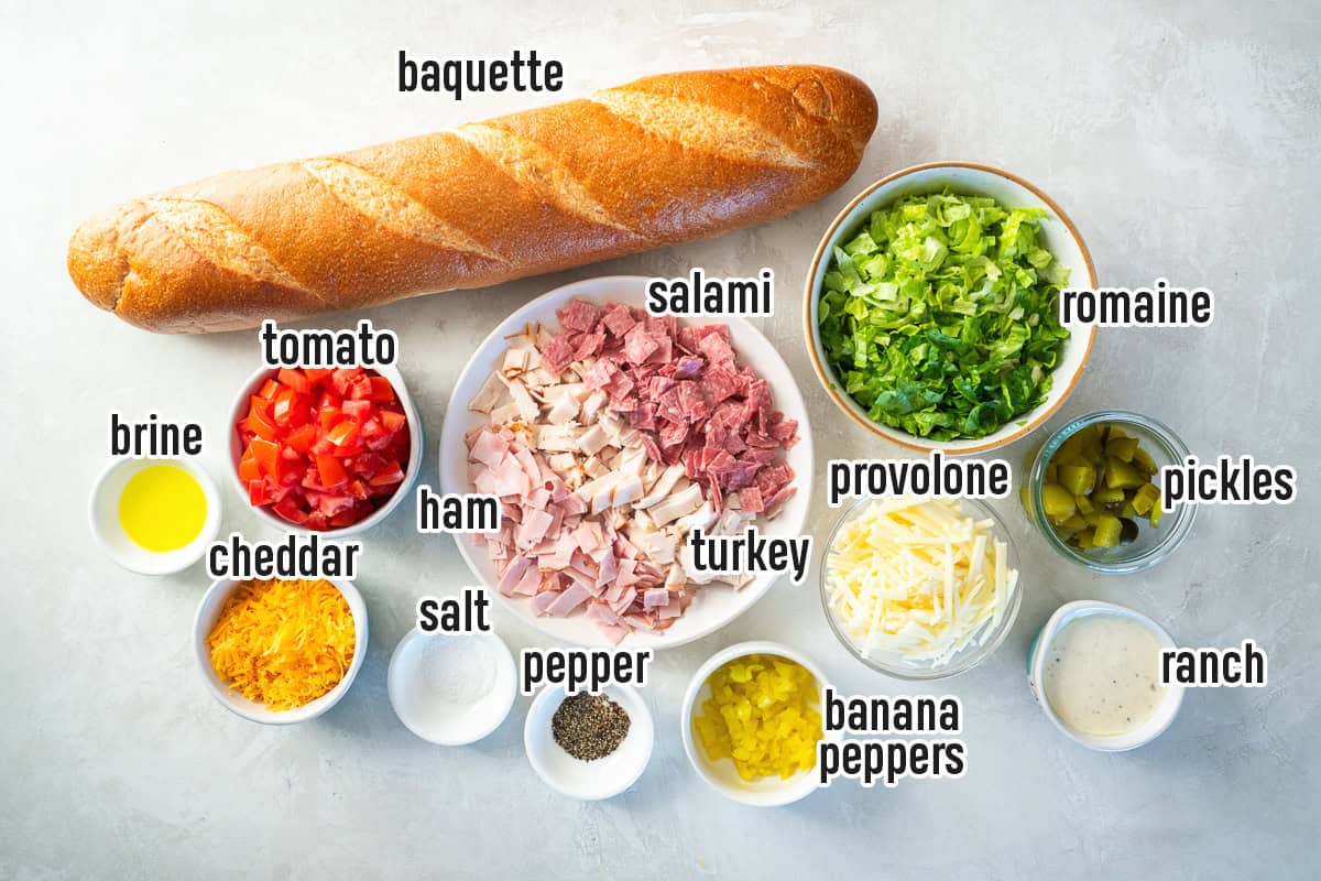 Chopped Italian Sandwich ingredients arranged on a light surface, including a baguette, chopped romaine, tomato, turkey, ham, salami, provolone, cheddar, banana peppers, pickles, ranch dressing, brine, salt, and pepper.