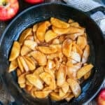 Cinnamon apples coated in a glossy brown sugar cinnamon sauce in a cast iron skillet, surrounded by fresh apples and cinnamon sticks.