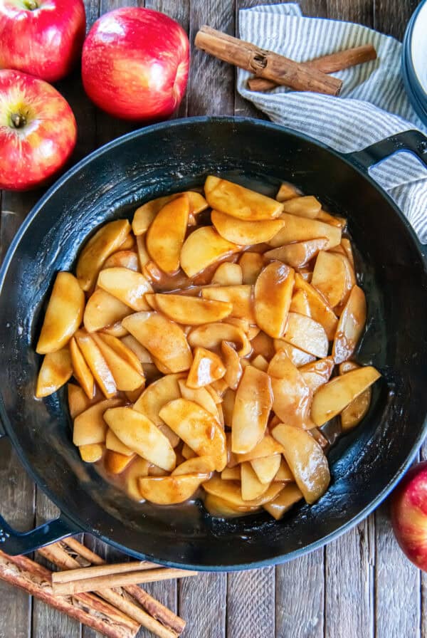 Cinnamon apples coated in a glossy brown sugar cinnamon sauce in a cast iron skillet, surrounded by fresh apples and cinnamon sticks.