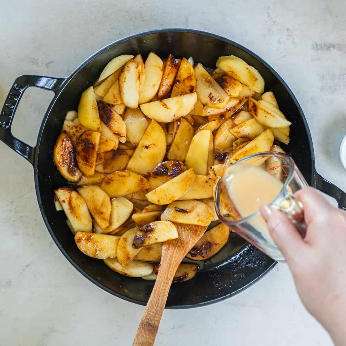 Apple juice and cornstarch mixture being poured over cinnamon apples in a skillet.