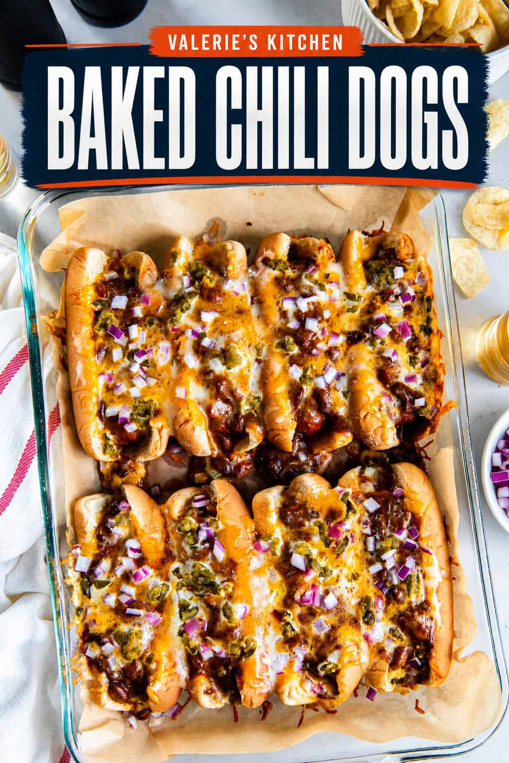 Eight baked chili dogs topped with melted cheese, red onion, and pickled jalapeños in a baking dish with beer and potato chips.