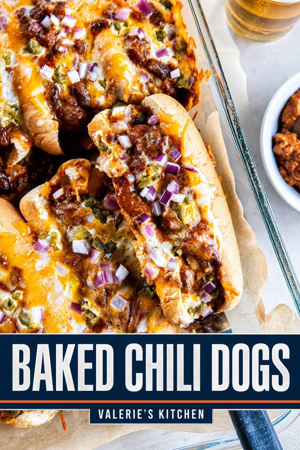 A baked chili dog being lifted from a pan of chili dogs topped with melted cheese, red onion, and pickled jalapeños with bold title text overlay.