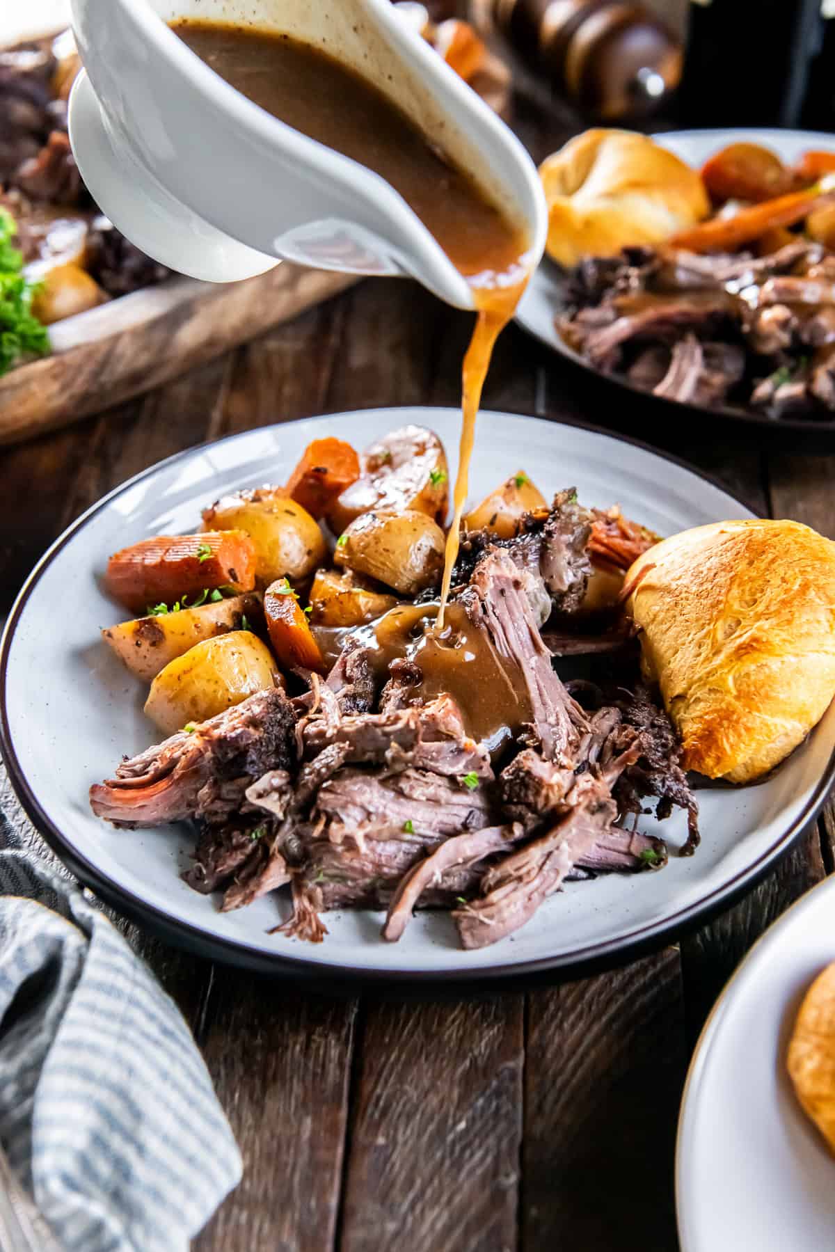 Brown gravy being poured over tender slow cooker chuck roast with carrots, potatoes, and a golden dinner roll.