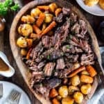 Tender slow cooker chuck roast with carrots and baby potatoes on a wooden serving platter, garnished with fresh parsley and served with gravy.
