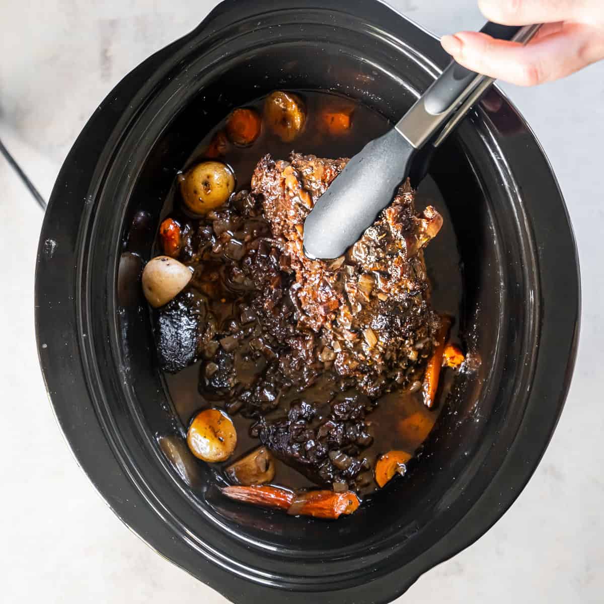 Tongs lifting a tender cooked chuck roast from a slow cooker.