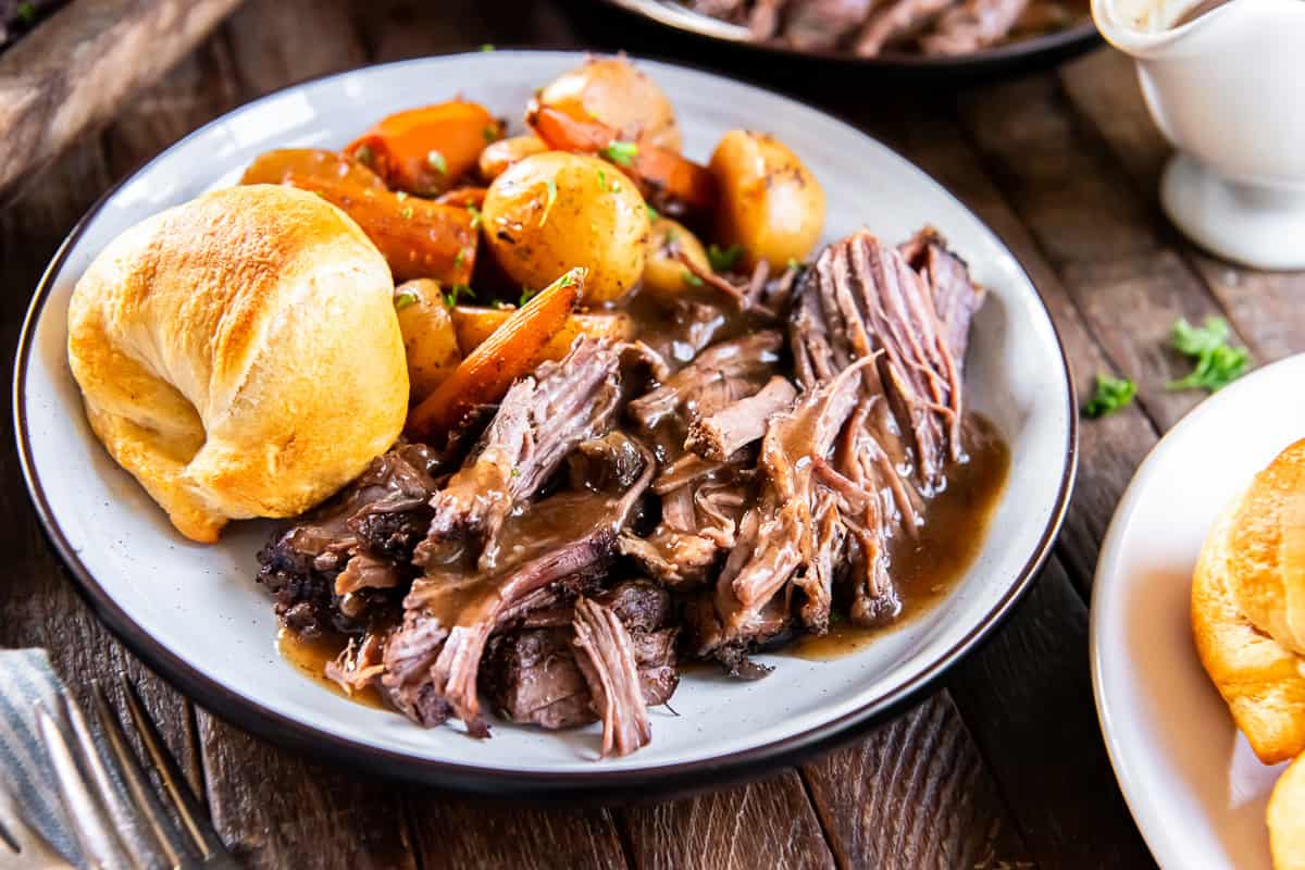 Tender slow cooker chuck roast with rich brown gravy, carrots, baby potatoes, and a golden dinner roll on a plate.