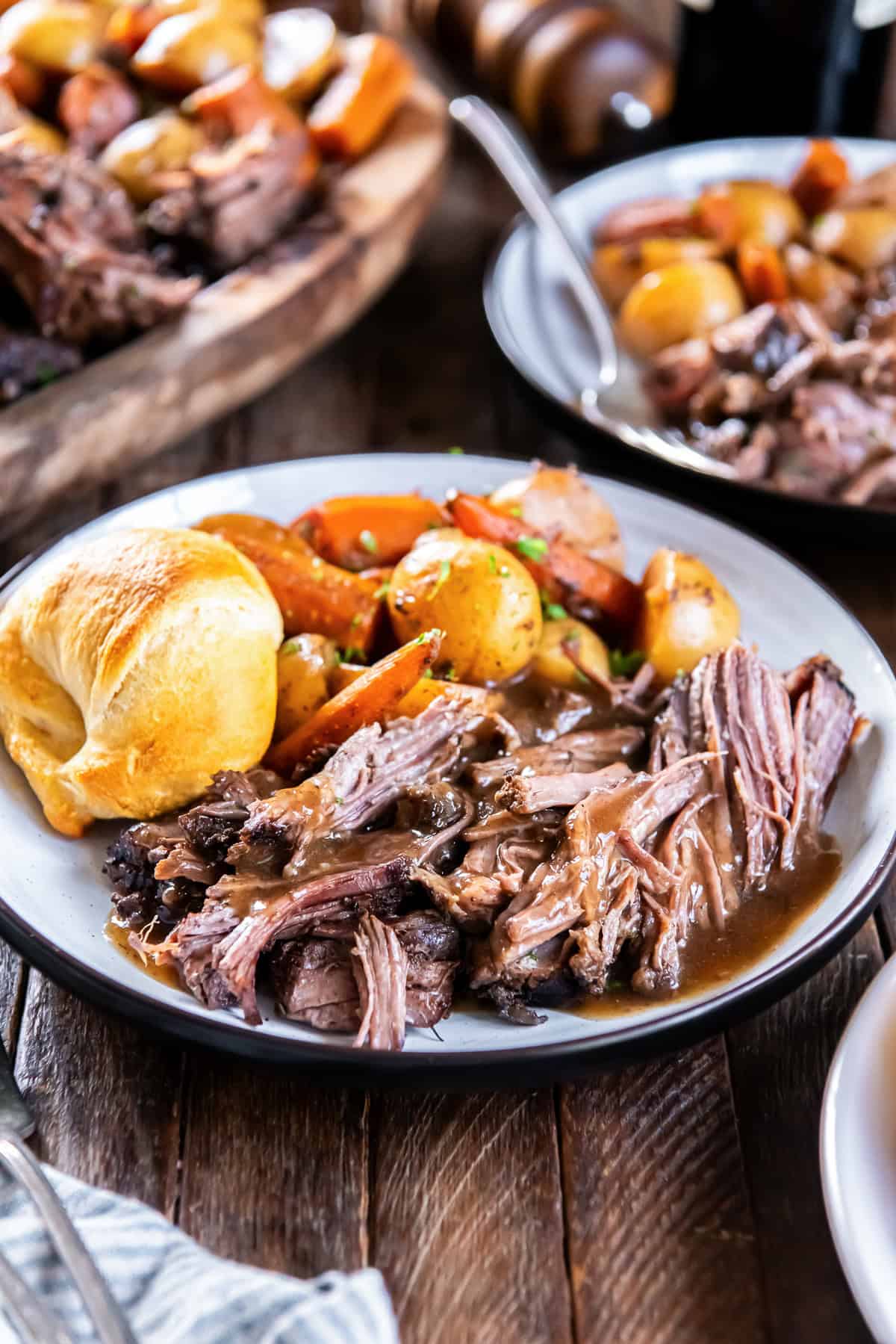 Tender slow cooker chuck roast with rich brown gravy, baby potatoes, carrots, and a golden dinner roll on a white plate.