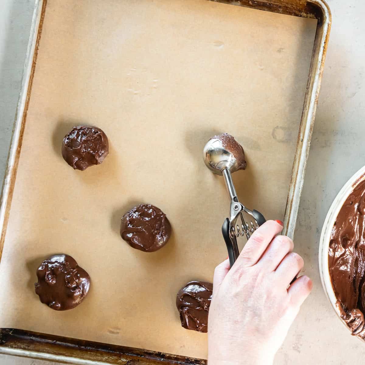 Chocolate brownie cookie dough being portioned onto a parchment-lined baking sheet with a cookie scoop.
