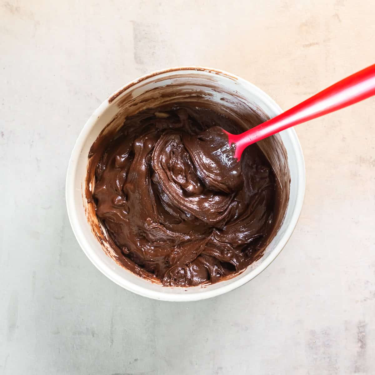 Glossy chocolate brownie cookie dough with chocolate chips in a white mixing bowl with a red spatula.