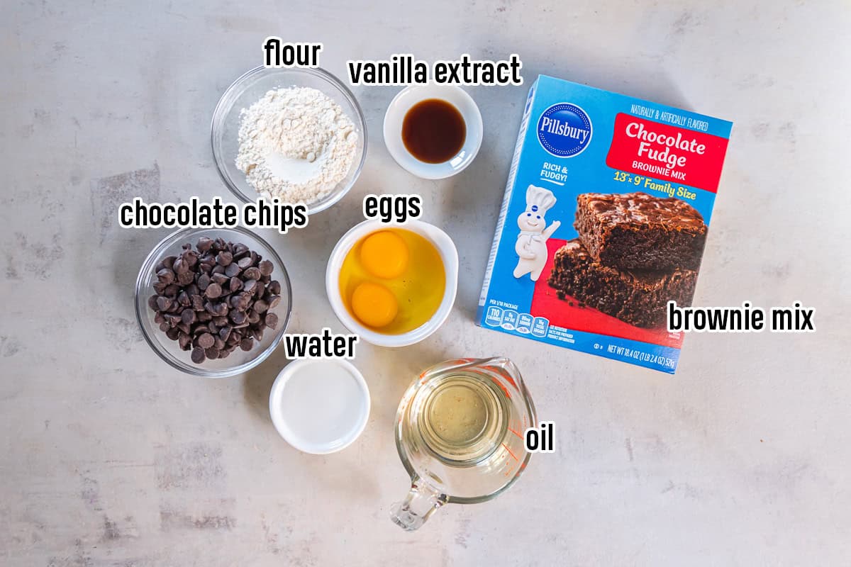 Ingredients for brownie mix cookies, including a box of Pillsbury Chocolate Fudge Brownie Mix, eggs, flour, vegetable oil, water, vanilla extract, and semisweet chocolate chips arranged in bowls and a measuring cup.