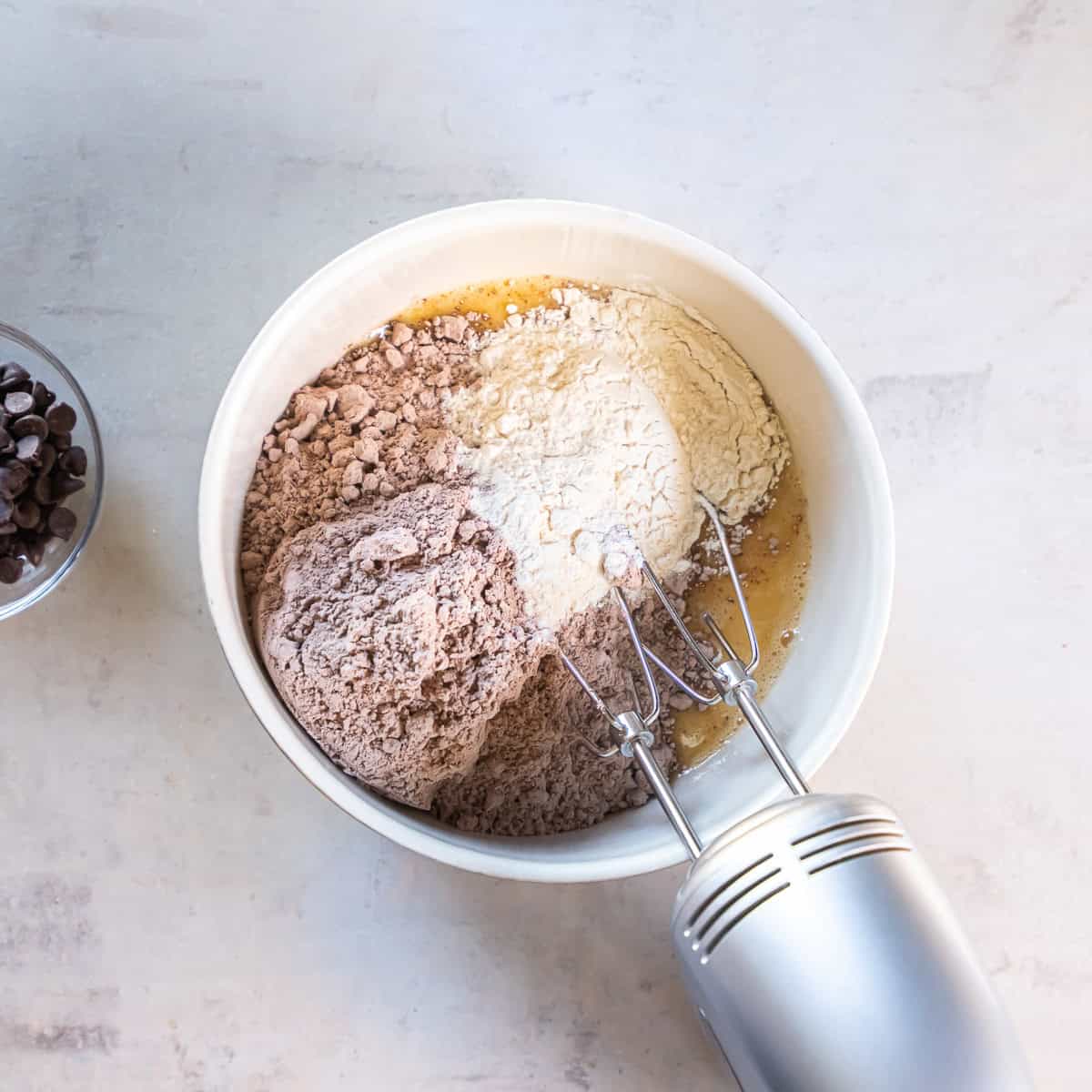 Brownie mix and all-purpose flour added to the wet ingredients in a white mixing bowl with an electric hand mixer.