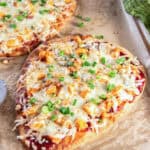 Buffalo chicken flatbread pizza topped with melted mozzarella, crumbled blue cheese, and chopped green onions on a parchment-lined baking sheet, with a second pizza behind it and a green towel at the side.