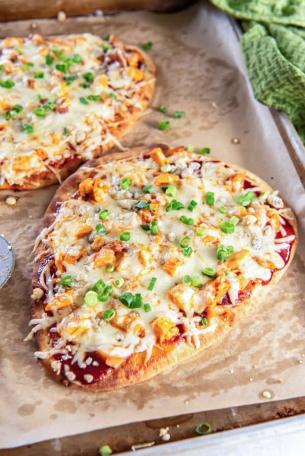 Buffalo chicken flatbread pizza topped with melted mozzarella, crumbled blue cheese, and chopped green onions on a parchment-lined baking sheet, with a second pizza behind it and a green towel at the side.