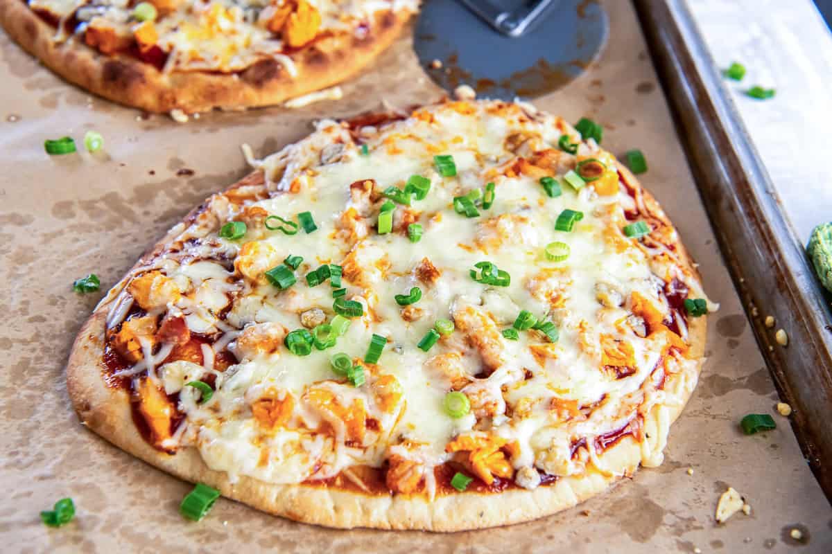 Buffalo chicken flatbread pizza topped with melted cheese and chopped green onions on a parchment-lined baking sheet, with a second pizza and pizza cutter behind it.