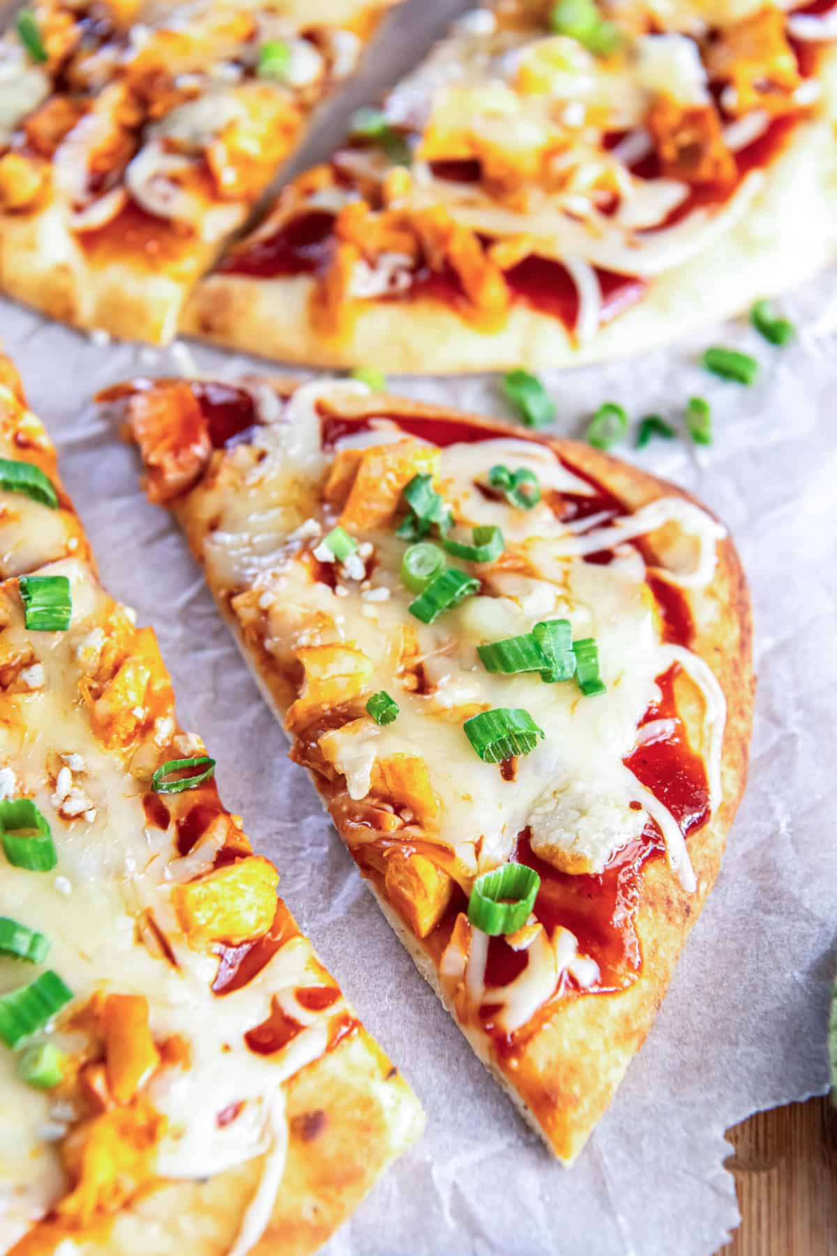 A slice of Buffalo chicken flatbread pizza topped with melted cheese and chopped green onions on parchment paper, with more pizza slices behind it.