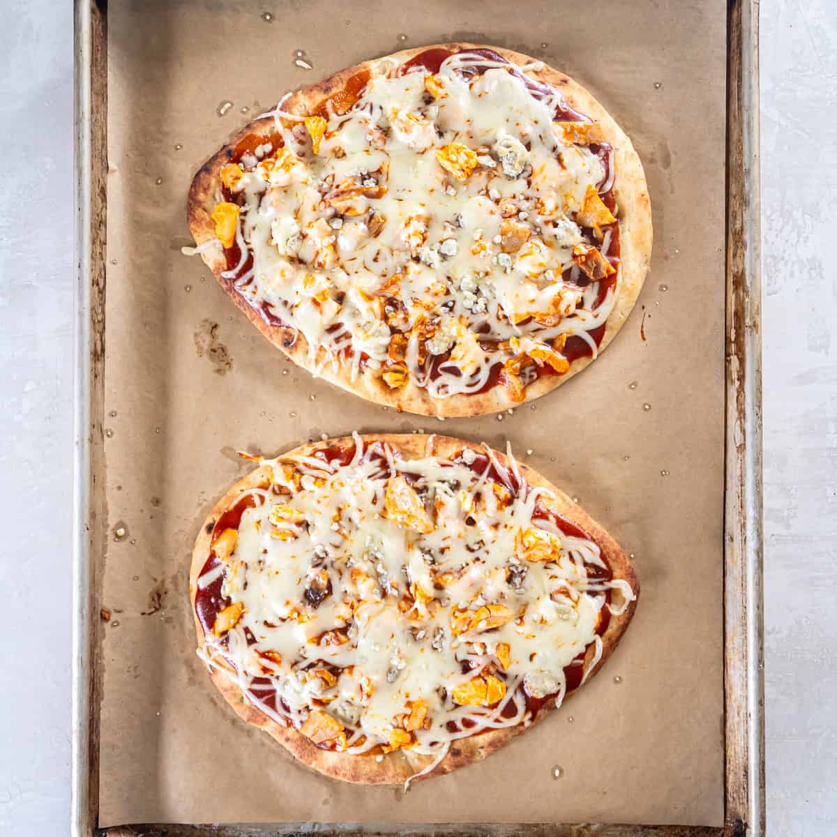 Two baked chicken flatbread pizzas with melted cheese and browned edges on a parchment-lined baking sheet.