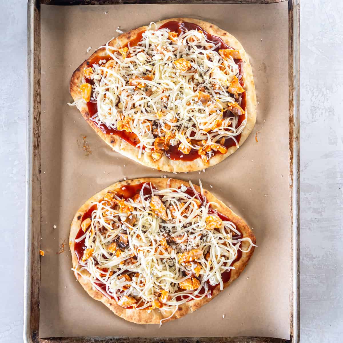 Two unbaked flatbread pizzas topped with sauce, chicken, shredded mozzarella, and crumbled blue cheese on a parchment-lined baking sheet.