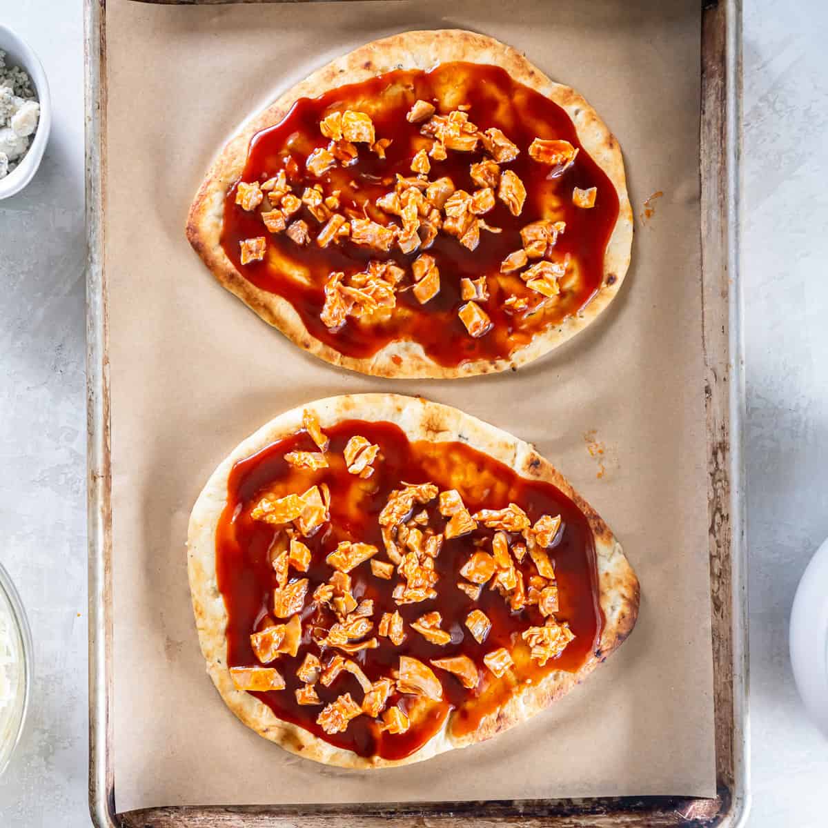 Two naan flatbreads topped with reddish-brown sauce and pieces of Buffalo chicken on a parchment-lined baking sheet.
