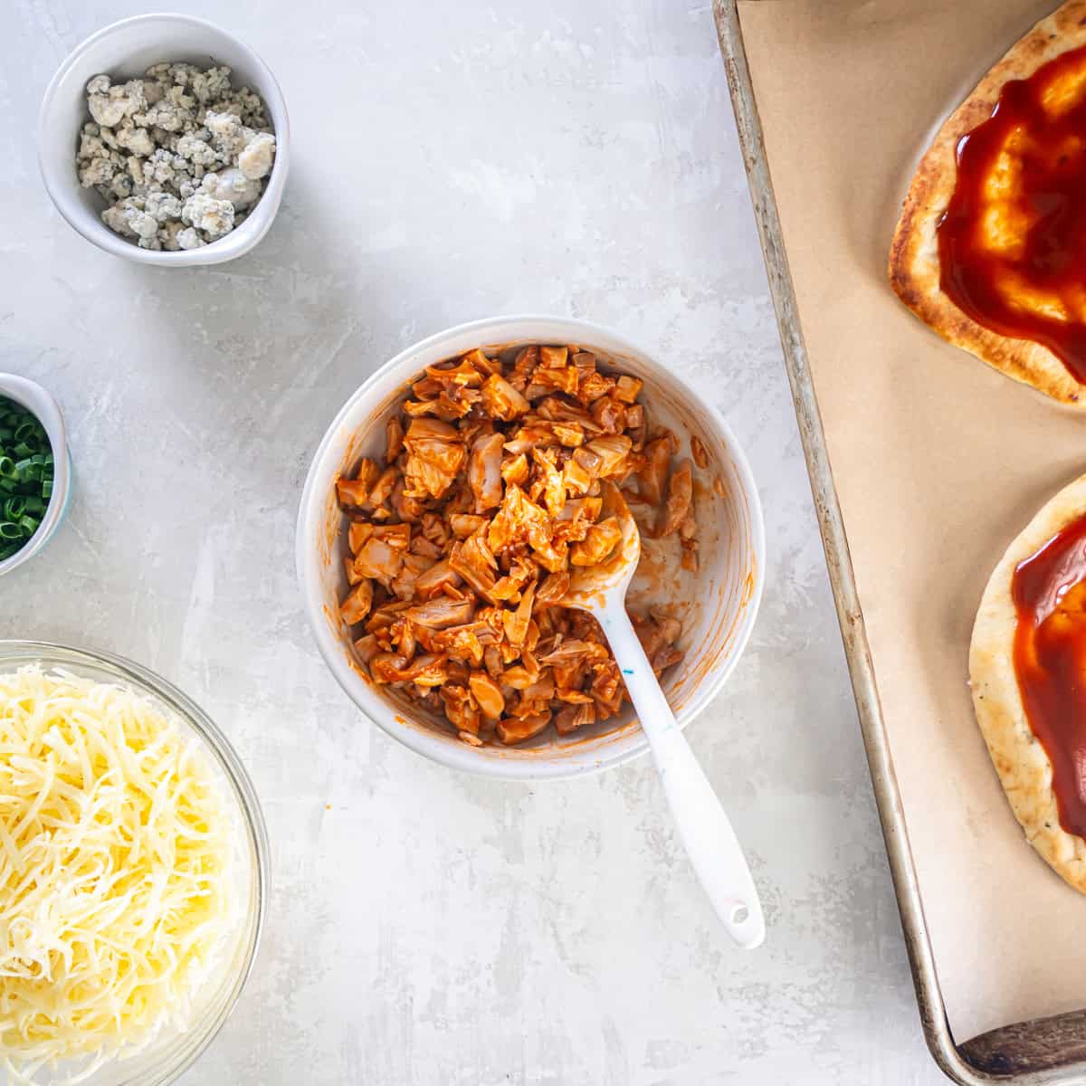 Buffalo sauce-coated chicken in a white bowl with a spatula, beside bowls of cheese and sauce-covered naan.