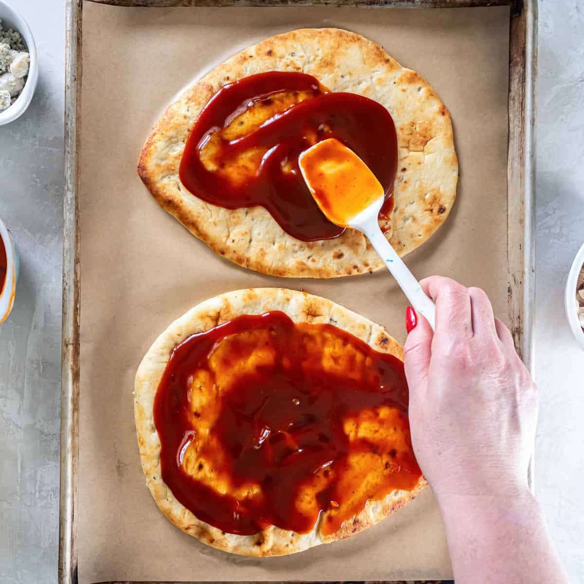 A hand spreading sauce over two naan breads with a white spatula on a parchment-lined baking sheet.