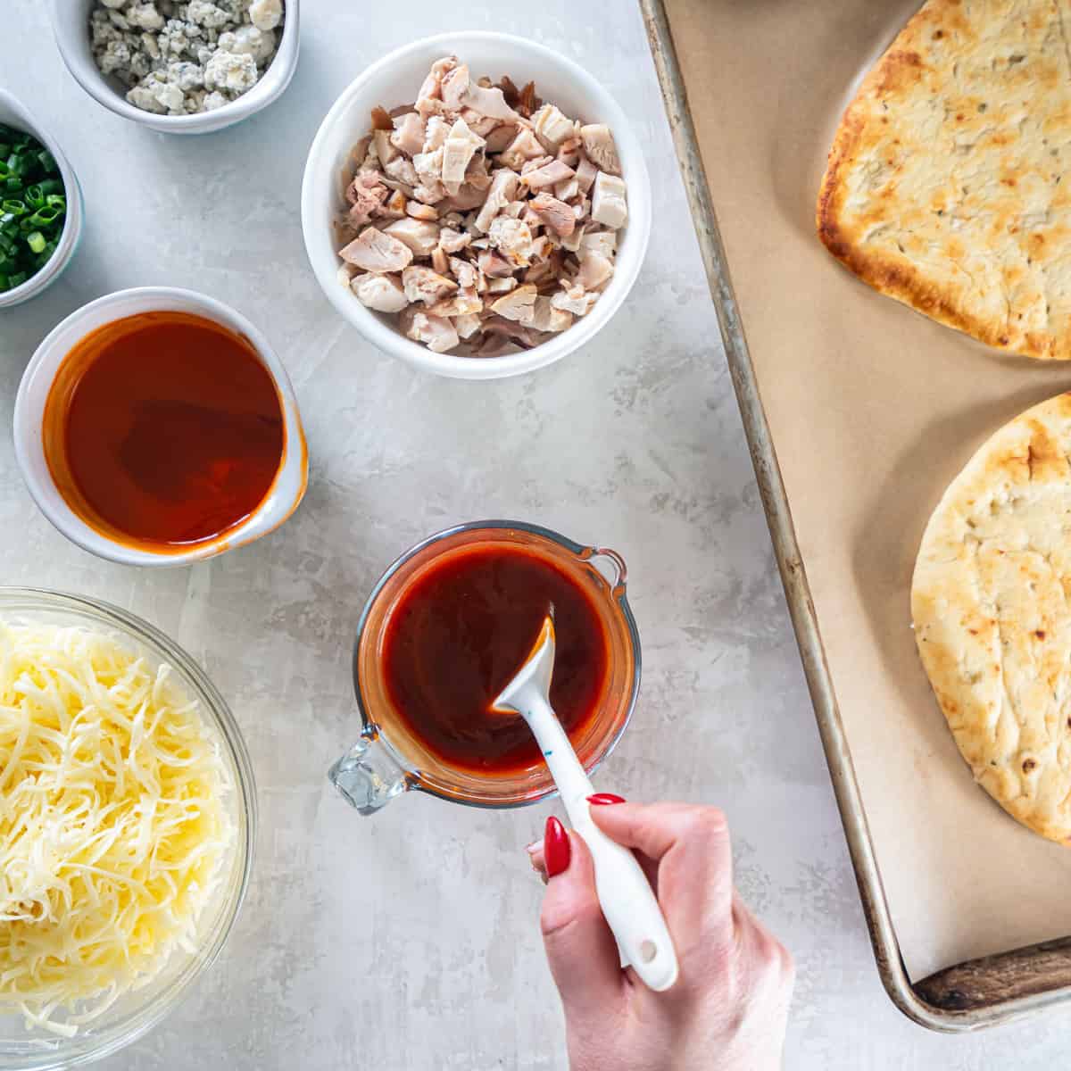 A hand stirring reddish-brown sauce in a glass measuring cup beside bowls of chicken and cheese, with plain naan on a lined baking sheet.