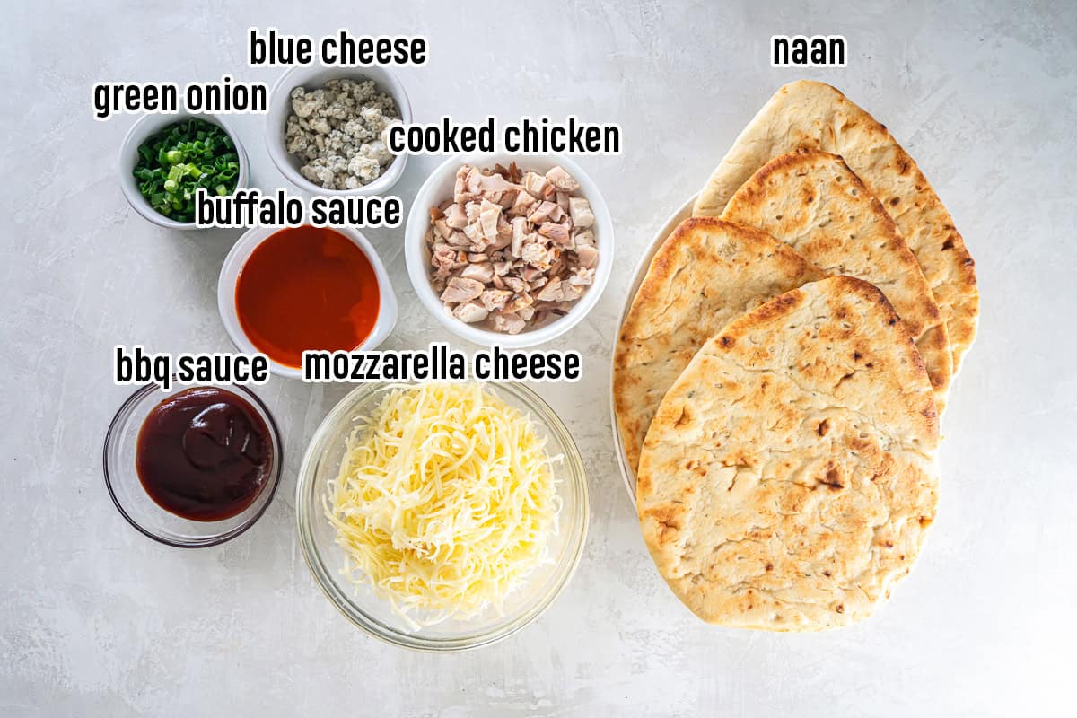 Four overlapping naan breads on a white platter beside bowls of cooked chicken, shredded mozzarella, crumbled blue cheese, chopped green onion, Buffalo sauce, and barbecue sauce, with each ingredient labeled.
